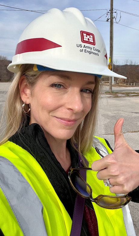 A woman in a white hardhat and a yellow safety vest gives a thumbs up.