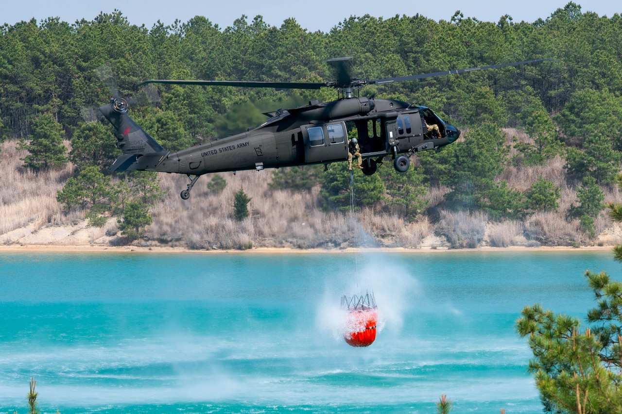 A Black Hawk helicopter hovers over water as it refills a bucket used for fighting wildfires.
