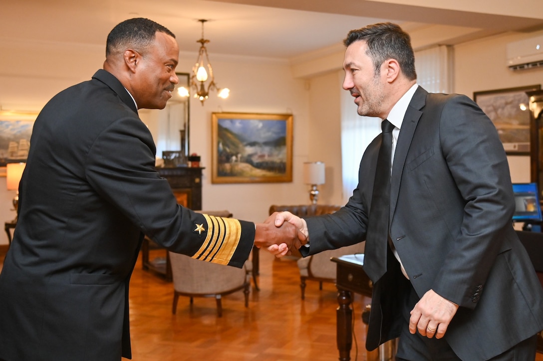 A man in a black military uniform shakes hand with a man in a dark gray suit.