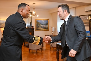 A man in a black military uniform shakes hand with a man in a dark gray suit.