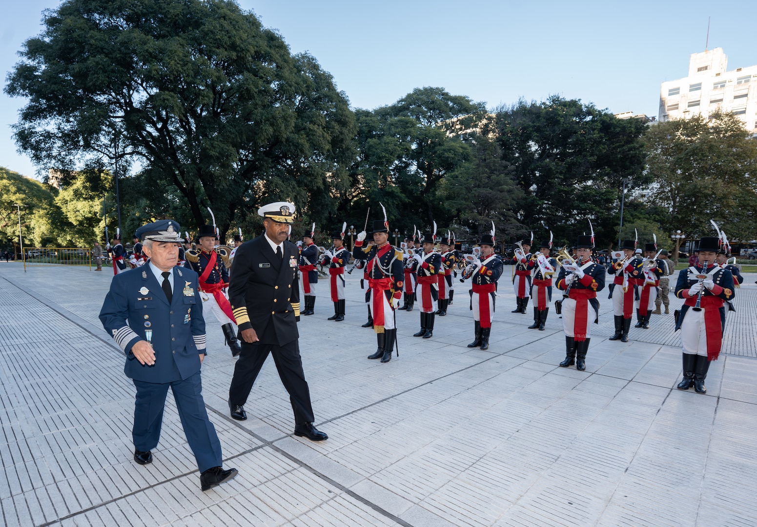 Two men in military dress uniforms walk with a formation of troops in the background.