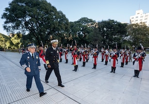 Two men in military dress uniforms walk with a formation of troops in the background.