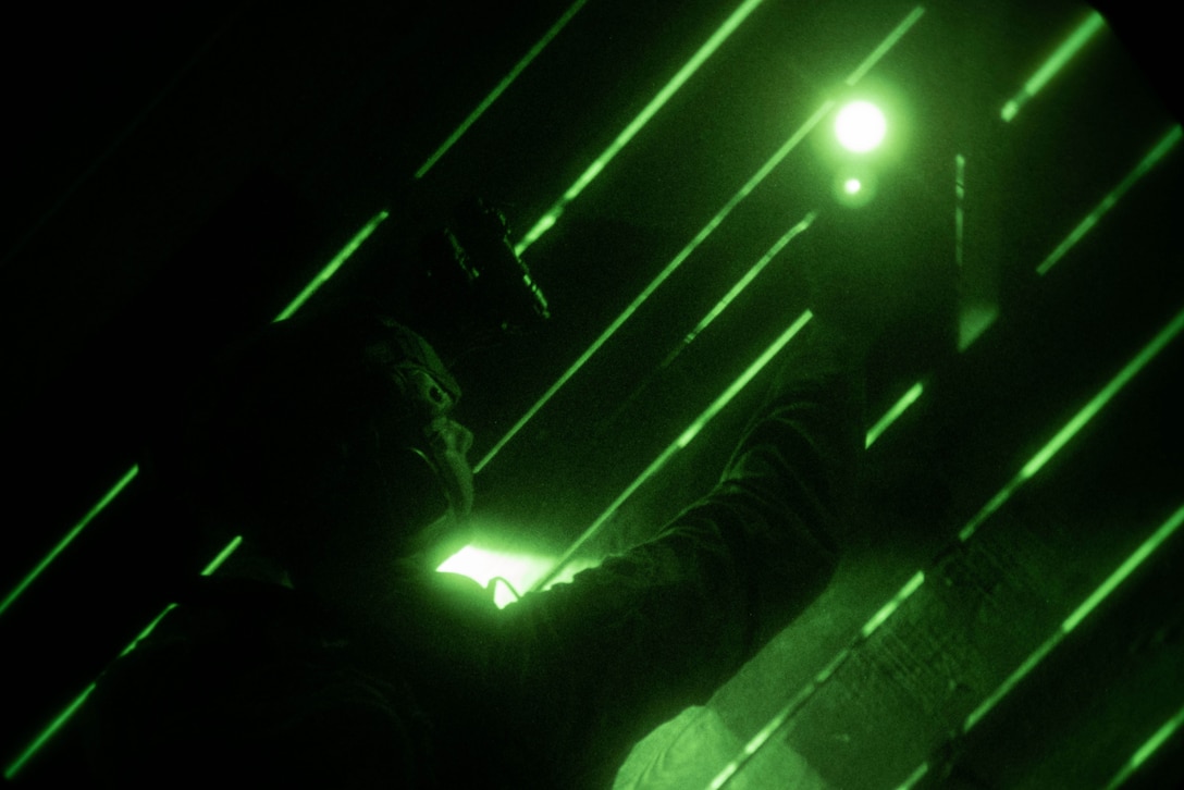Via night vision lens, a Marine stands holding a light up with one arm against diagonal rays of green light.