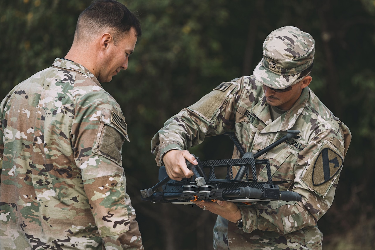 A soldier holds a drone outdoors while another looks at it.