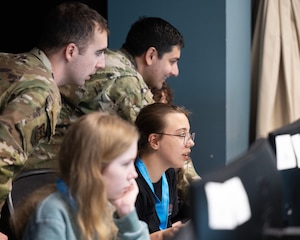 Two teenage girls sit at computers withe two men in military uniform stand behind them. All are looking intently at screens.