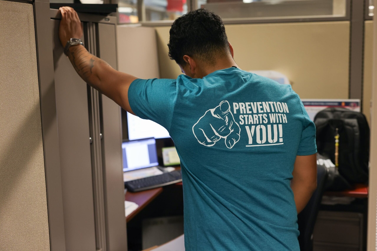 A man with his back to the camera is leaning against a filing cabinet while wearing a shirt that reads, “Prevention starts with you!”