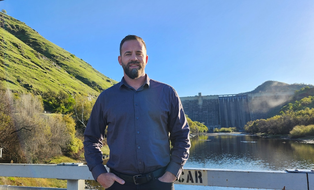 A man in a blue long-sleeved, button-up shirt stands with his hands in his pockets in front of a lake with Pine Flat Dam in the distance behind him.
