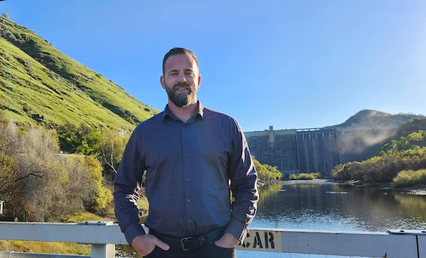 A man in a blue long-sleeved, button-up shirt stands with his hands in his pockets in front of a lake with Pine Flat Dam in the distance behind him.
