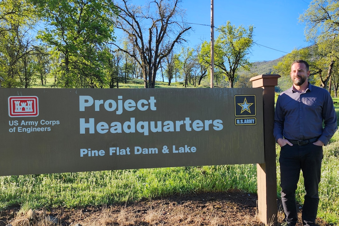 A man in blue, long-sleeved button-up shirt and jeans stands with his hands in his pants pockets to the right of a Pine Flat Dam and Lake sign that reads "Project Headquarters" and has the U.S. Army Corps of Engineers castle emblem and U.S. Army emblem on it.