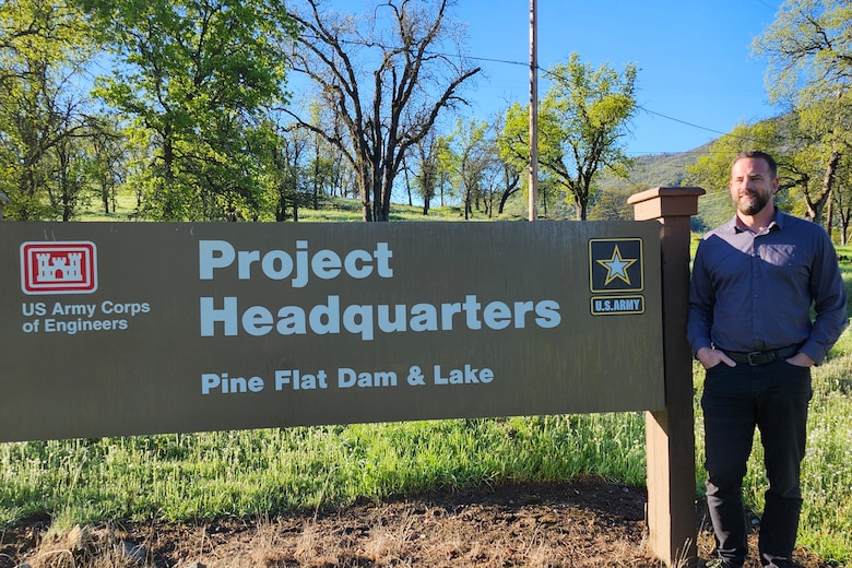 A man in blue, long-sleeved button-up shirt and jeans stands with his hands in his pants pockets to the right of a Pine Flat Dam and Lake sign that reads "Project Headquarters" and has the U.S. Army Corps of Engineers castle emblem and U.S. Army emblem on it.