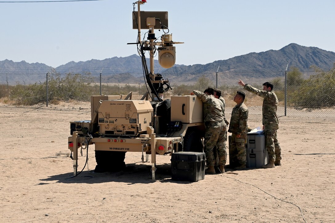 Four military police work on a military surveillance system on a sunny day with mountains in the distance.