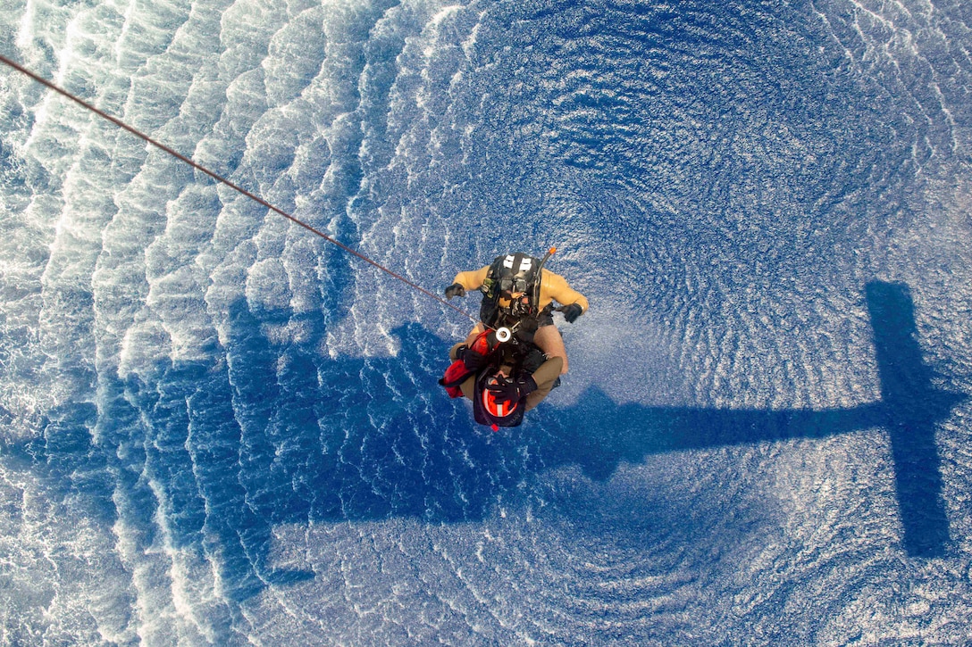 The shadow of a helicopter is seen on a body of water as two sailors attached to a line hover above it during daylight.
