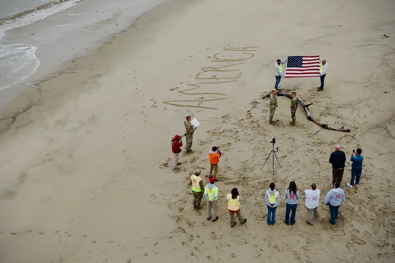 A dozen people stand near or behind a camera as it films two soldiers raising their right hands while facing each other on the beach in front of an American flag.