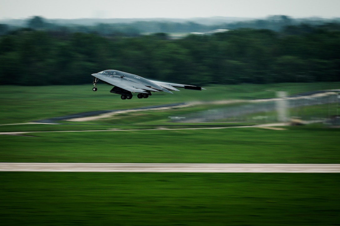 An aircraft takes off above a runway in a green field with a forest in the distance.