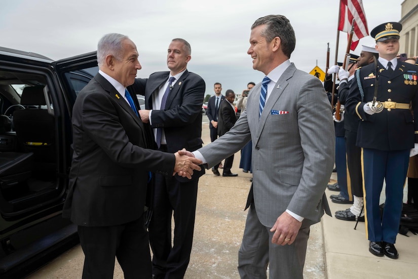 Two men in business attire shake hands as a man behind them in business attire holds a car door open. Service members in dress uniforms stand behind them.