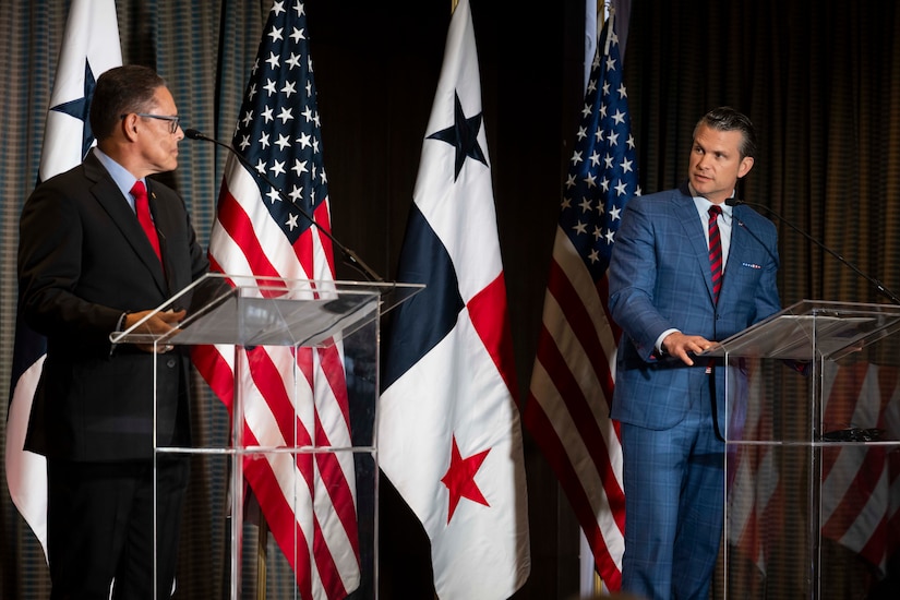 Two men in business attire, each behind a lectern, address each other. Behind them are the flags of the U.S. and Panama.