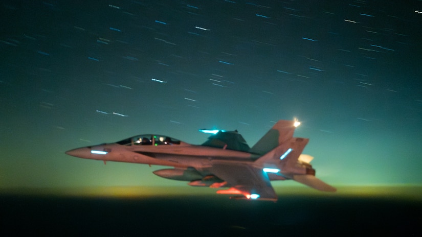 A fighter aircraft moves through the night sky and is lit up with a red light.