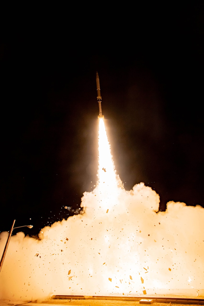 A missile is climbing upward through a dark sky with fire and smoke trailing behind it.