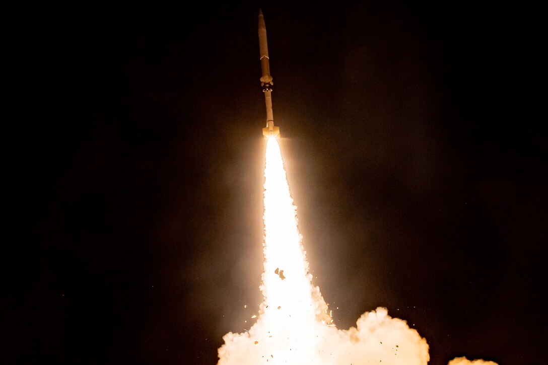 A missile is climbing upward through a dark sky with fire and smoke trailing behind it.