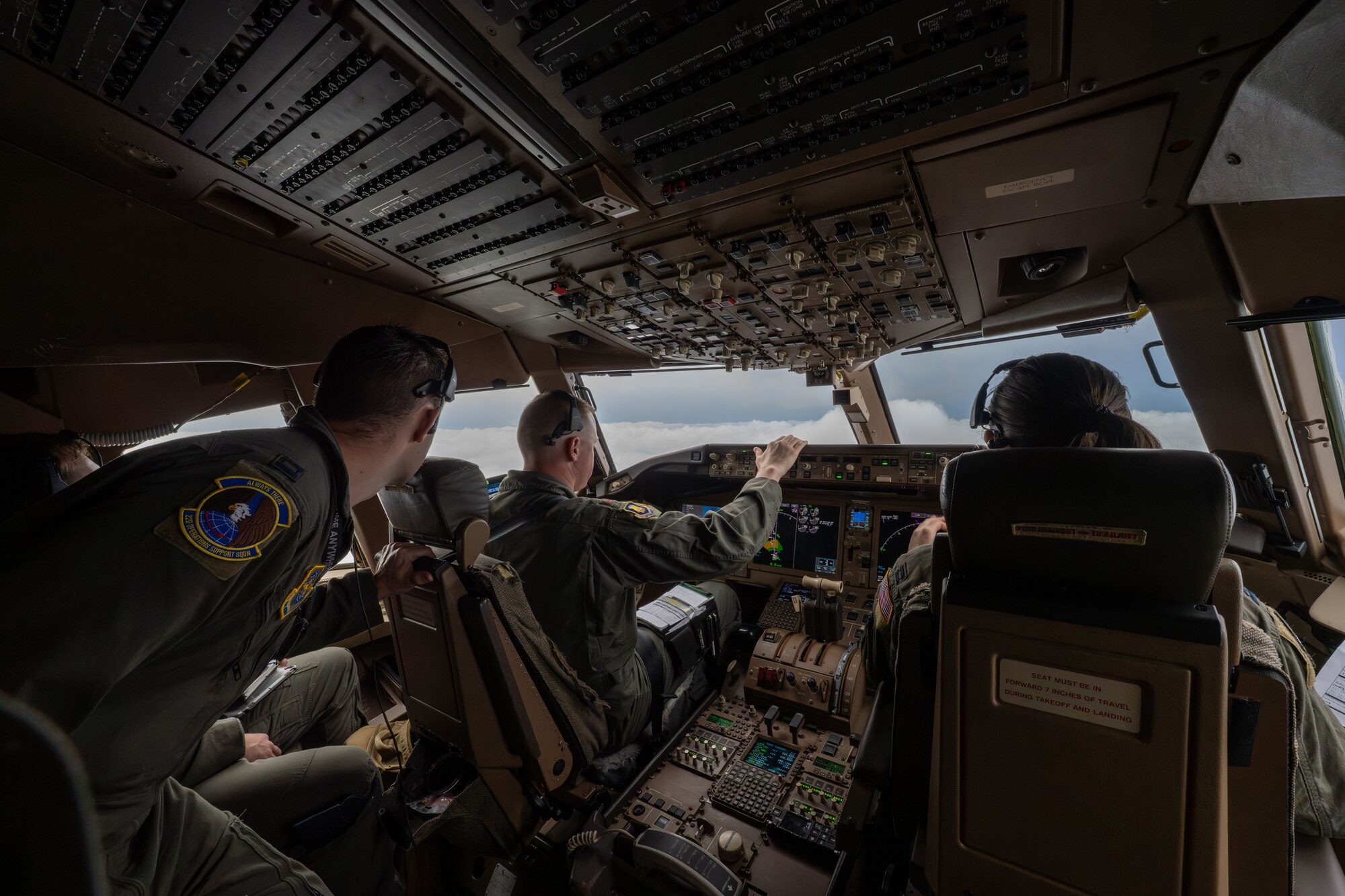 Three air crew personnel operate controls inside a KC-46 cockpit looking out into the clouds.
