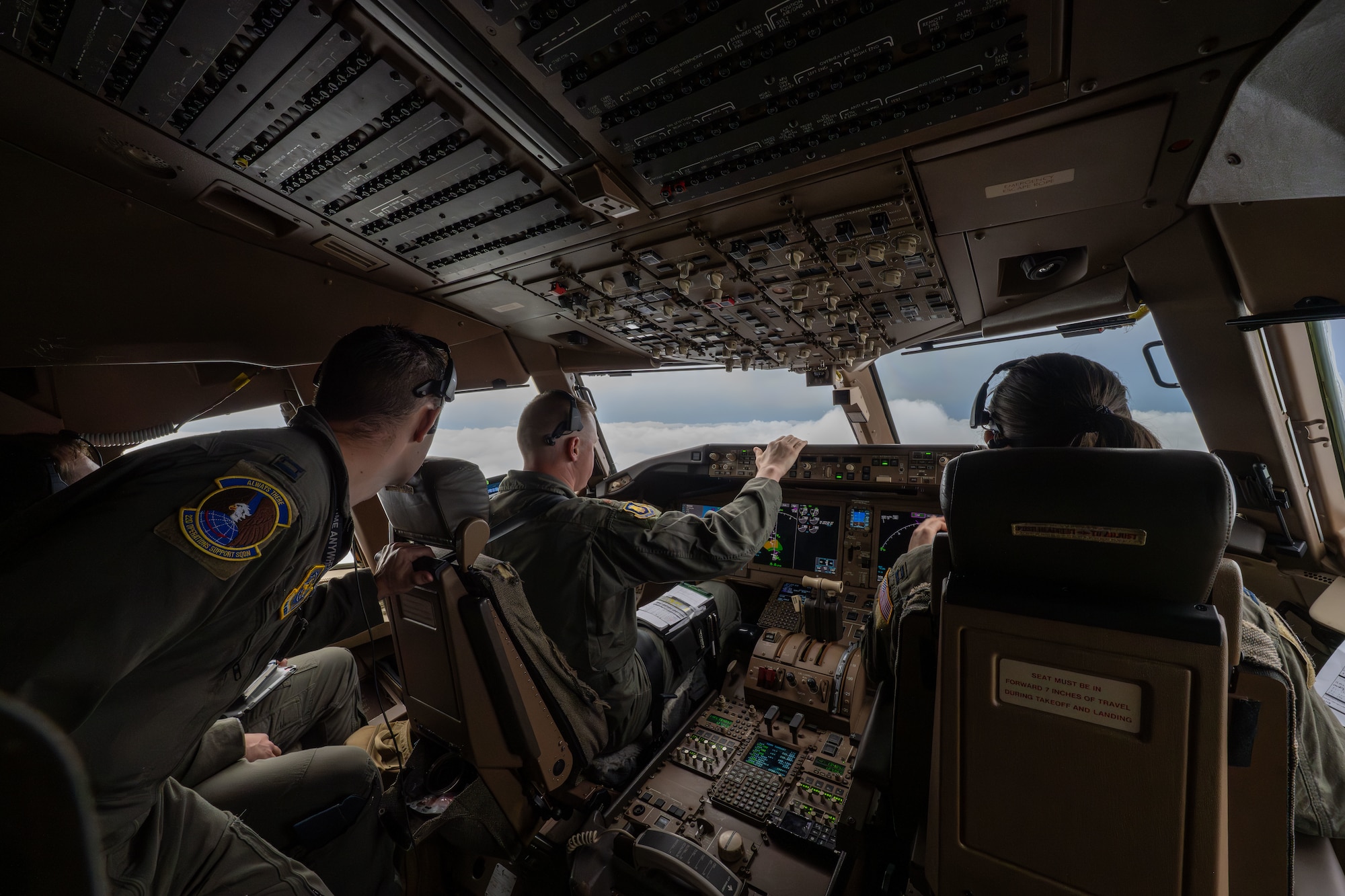 Three air crew personnel operate controls inside a KC-46 cockpit looking out into the clouds.