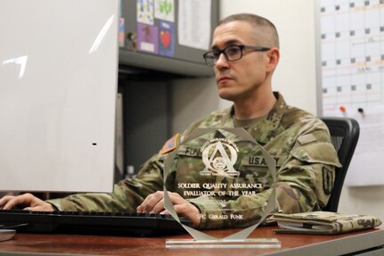 U.S. Army 1st Sgt. Gerald Funk, chief instructor for 2nd Battalion, 166th Regiment – Regional Training Institute, Pennsylvania Army National Guard, works at his office desk while his quality assurance evaluator of the year award, which he received during a U.S. Army quality assurance program forum at Fort Eustis, Va., sits on display April 30, 2025. (U.S. Army National Guard photo by Sgt. 1st Class Shane Smith)