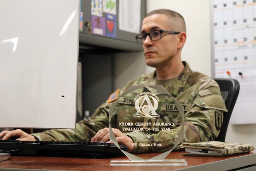 U.S. Army 1st Sgt. Gerald Funk, chief instructor for 2nd Battalion, 166th Regiment – Regional Training Institute, Pennsylvania Army National Guard, works at his office desk while his quality assurance evaluator of the year award, which he received during a U.S. Army quality assurance program forum at Fort Eustis, Va., sits on display April 30, 2025. (U.S. Army National Guard photo by Sgt. 1st Class Shane Smith)