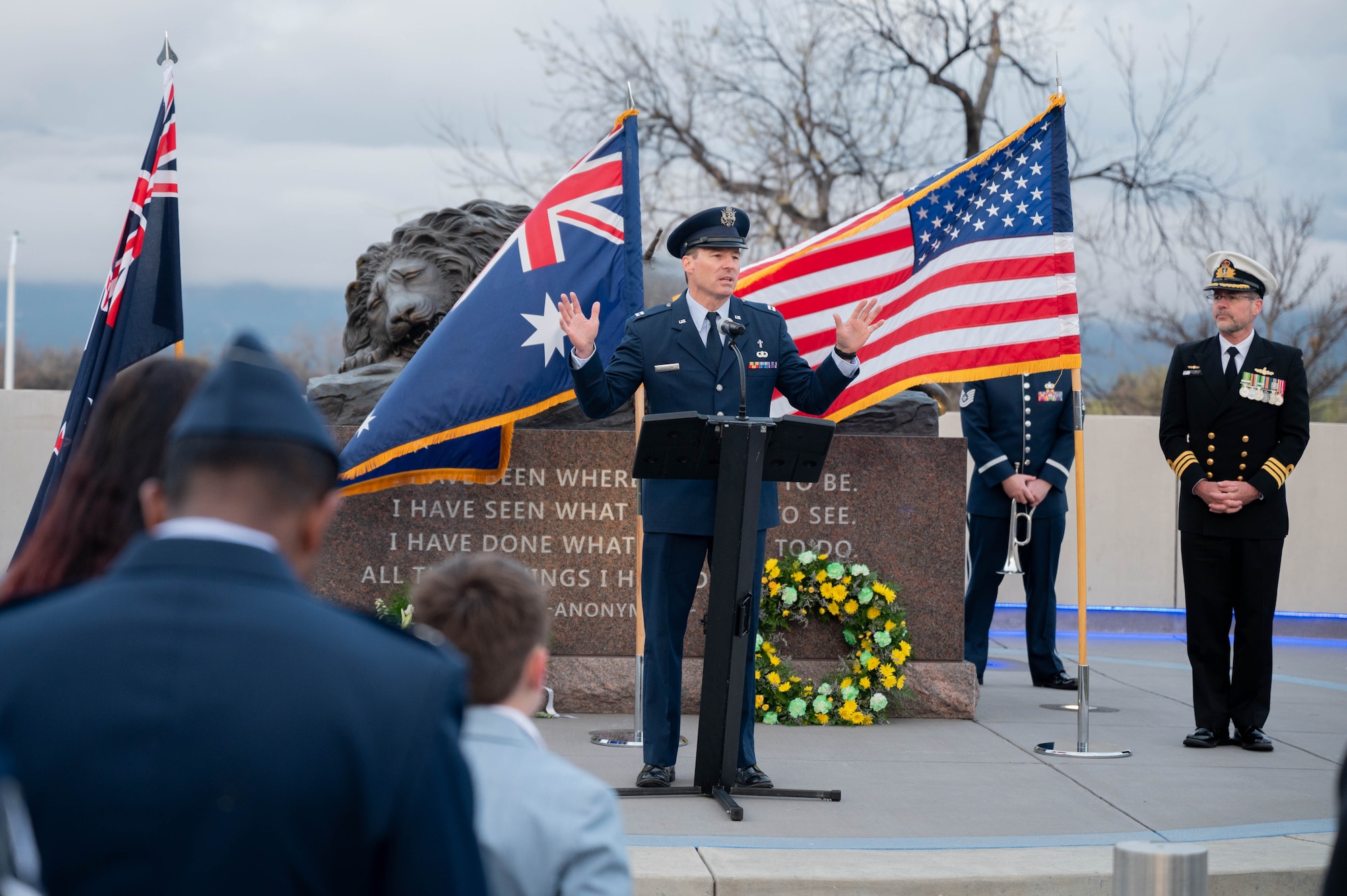 A man speaks to a crowd in front of a memorial with the American, Australian and New Zealand flags waving in the background.