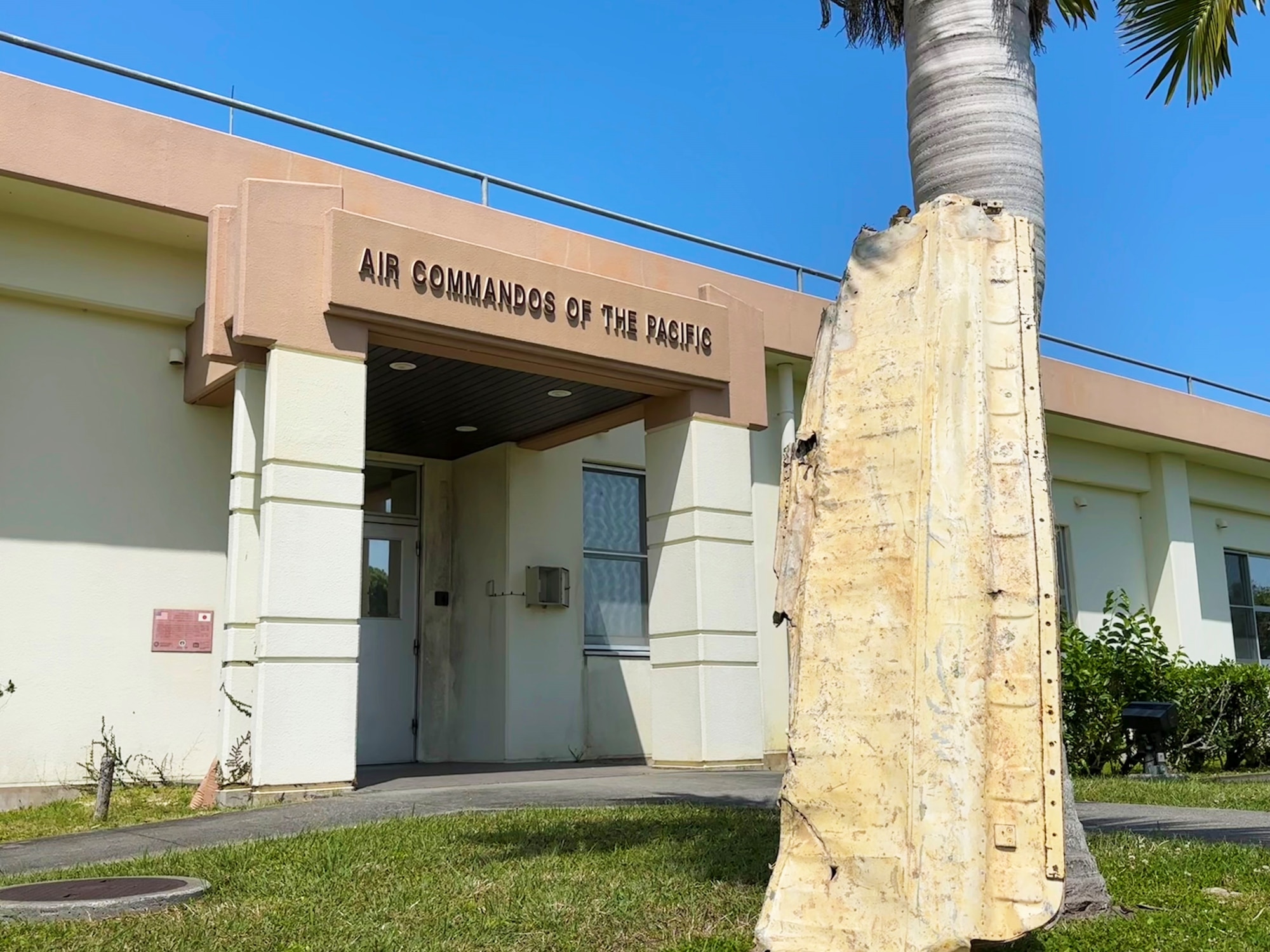 An old airplane wing rests against a tree in front of a building with the words "AIR COMMANDOS OF THE PACIFIC" above the door.