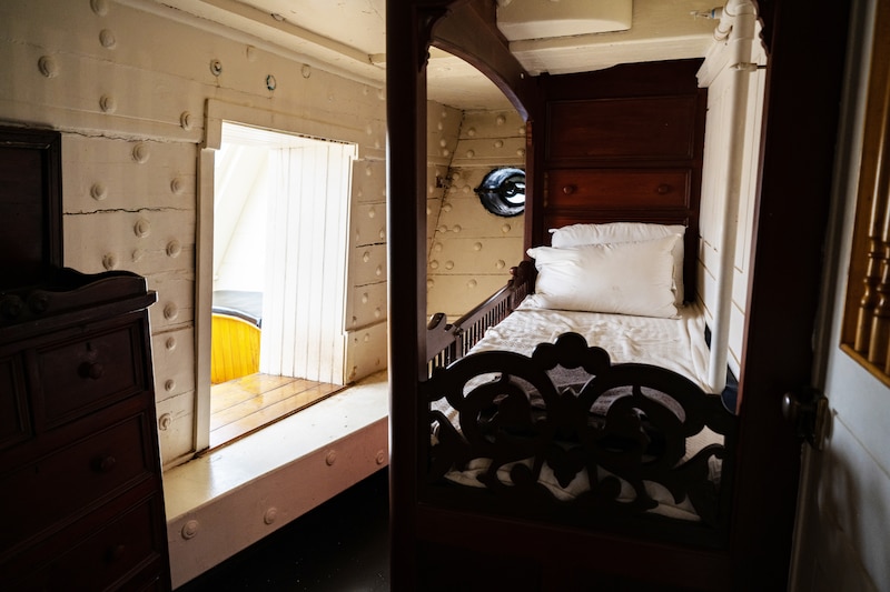 A wooden bed stands next to a window in a ship's cabin.