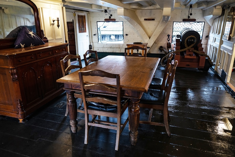A wooden desk and chairs are shown in a white cabin inside an old ship.