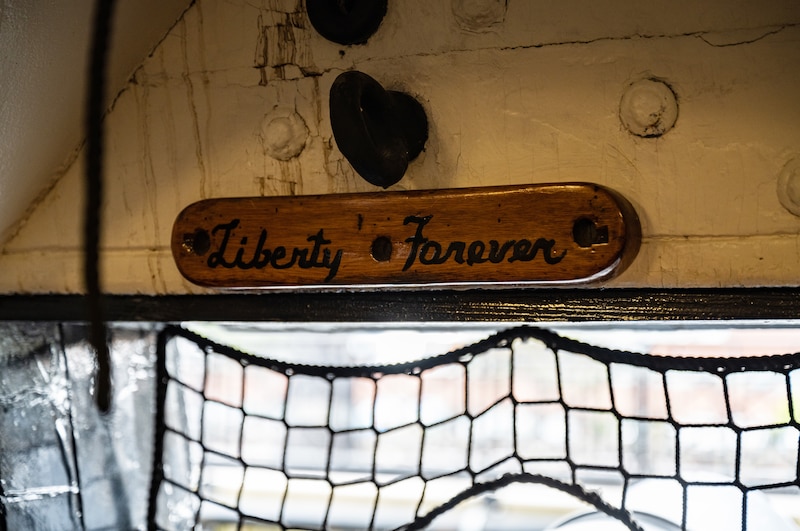 A nameplate with Liberty Forever written in cursive hangs above a window under a ship's deck.