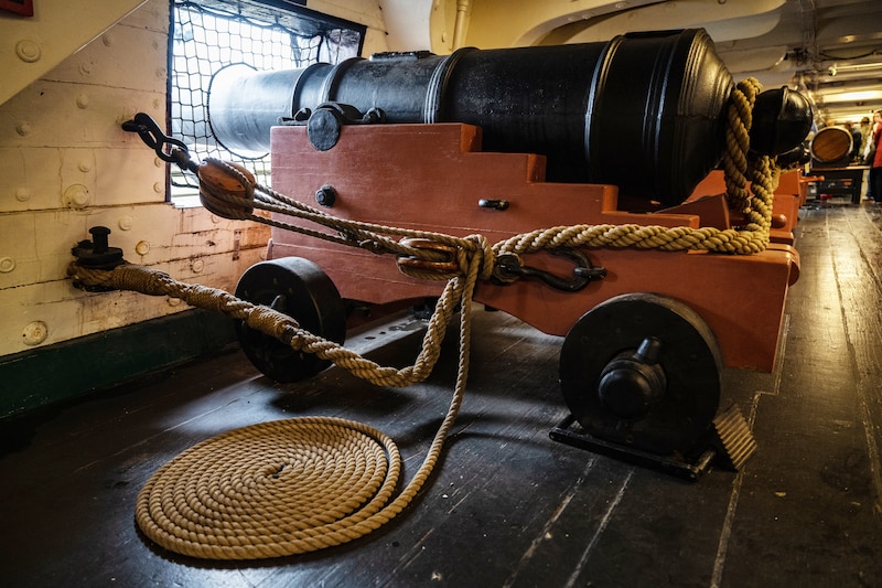 A canon on wheels is positioned inside of an old ship with rope next to it.