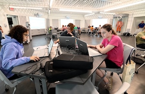 Classroom setting with three students sitting at the table in the forefront working on laptops; more students are the in background at tables with laptops