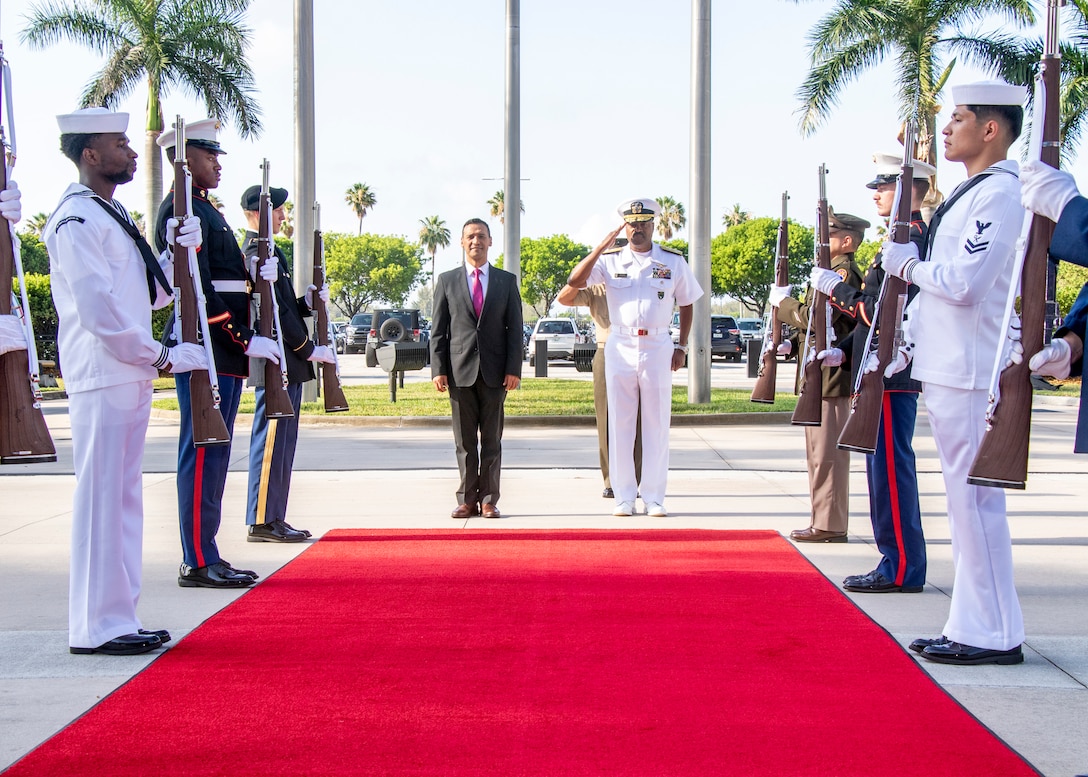 A man in a dark suit stands alongside a man in a white uniform saluting.