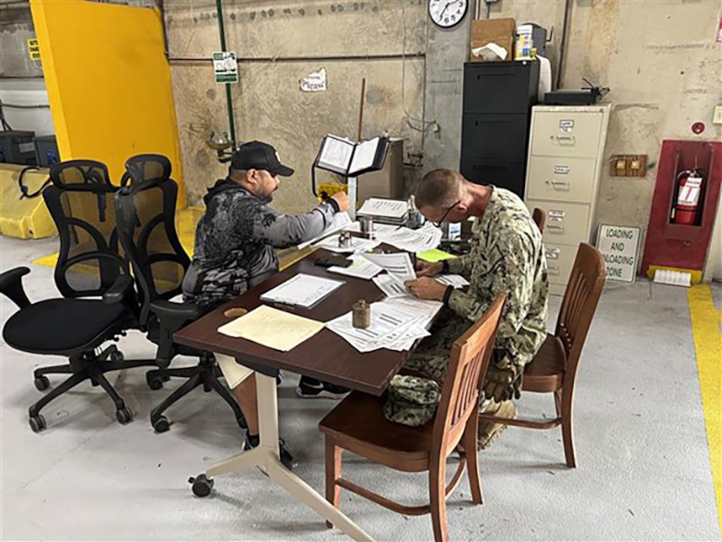 Two men look at paperwork while sitting at a warehouse desk.