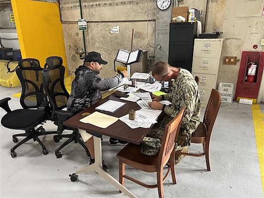 Two men look at paperwork while sitting at a warehouse desk.