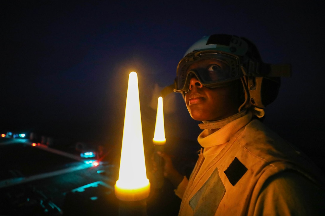 A sailor wearing goggles and holding two light signals looks out from the deck of a ship at night.