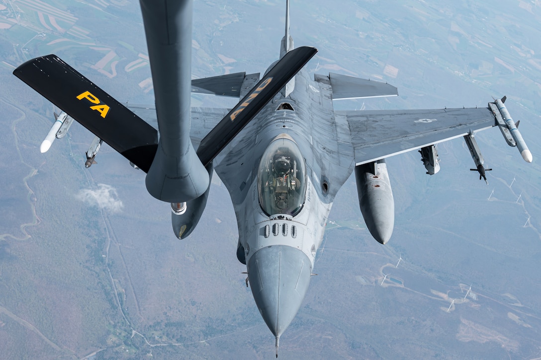 An airplane flies above a fighter jet during in-flight refueling training, with the ground seen below.
