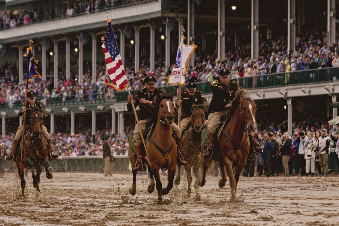 Four soldiers in uniform ride horses on a muddy racetrack as hundreds look on from a grandstand.