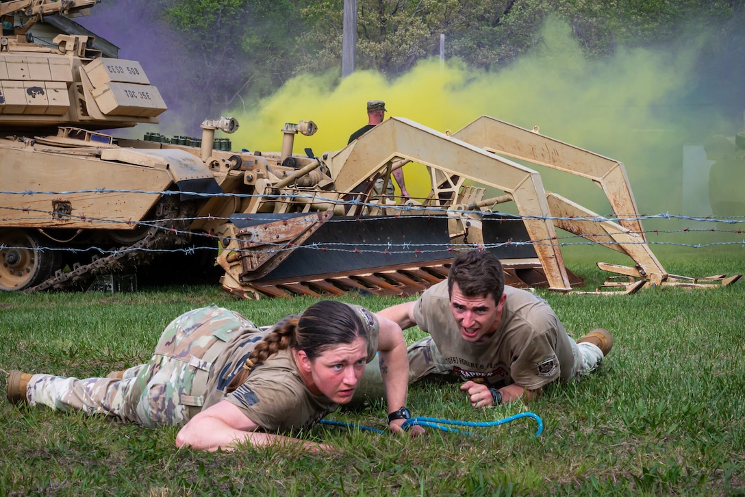 Two soldiers, one holding a rope, crawl under barbed wire with a tracked vehicle and yellow smoke in the background.