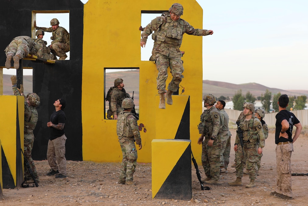 Soldiers move through and jump down from a yellow and black wall as others observe.