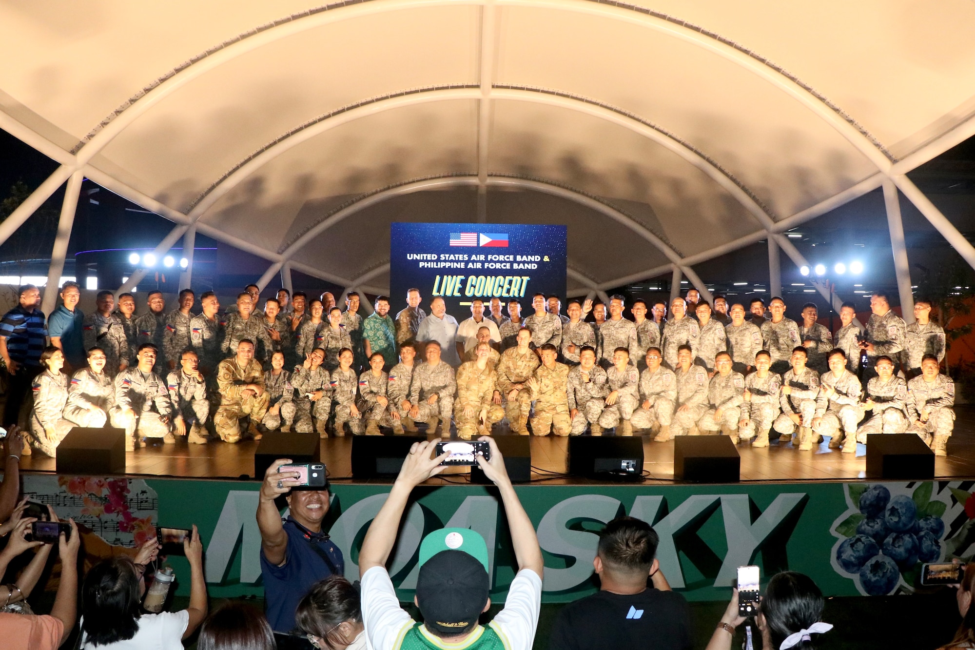 A large group of musicians in military uniforms pose on a concert stage as concert attendees take photos of them.