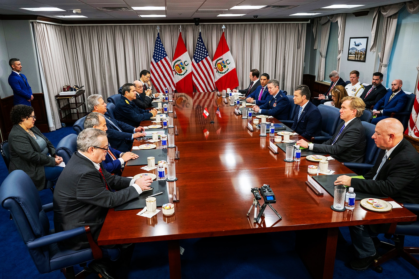A group of people sit at a long table with standing flags in the background.