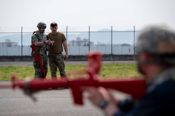 In the background a military member speaks to another military member and in the foreground a military member holds a red training weapon.