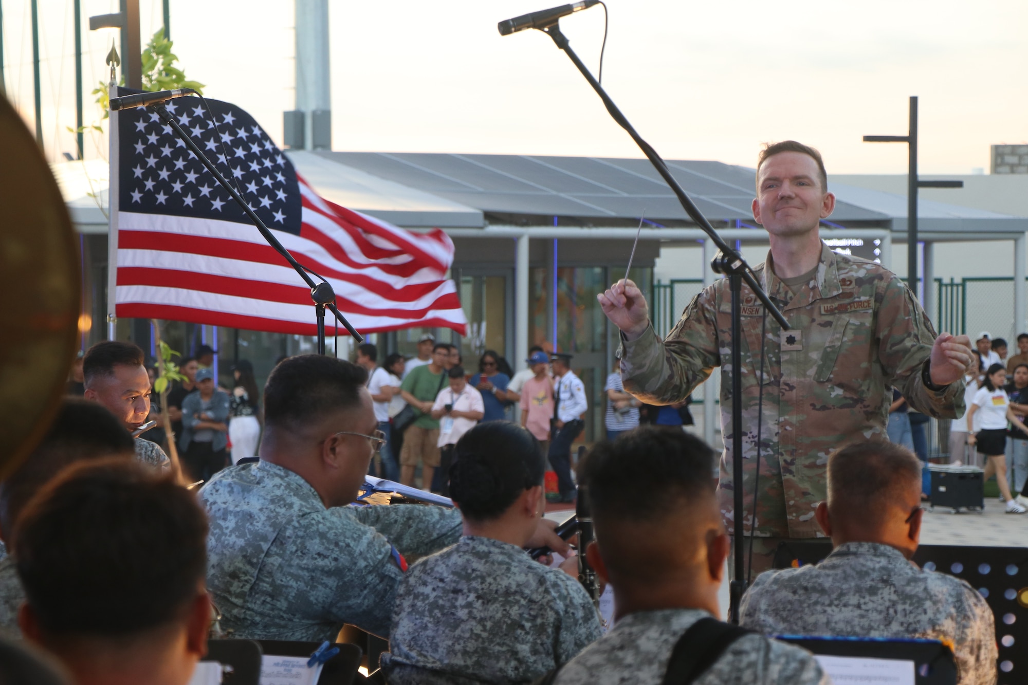 A soldier conducts a large concert band as a US flag waves in the background