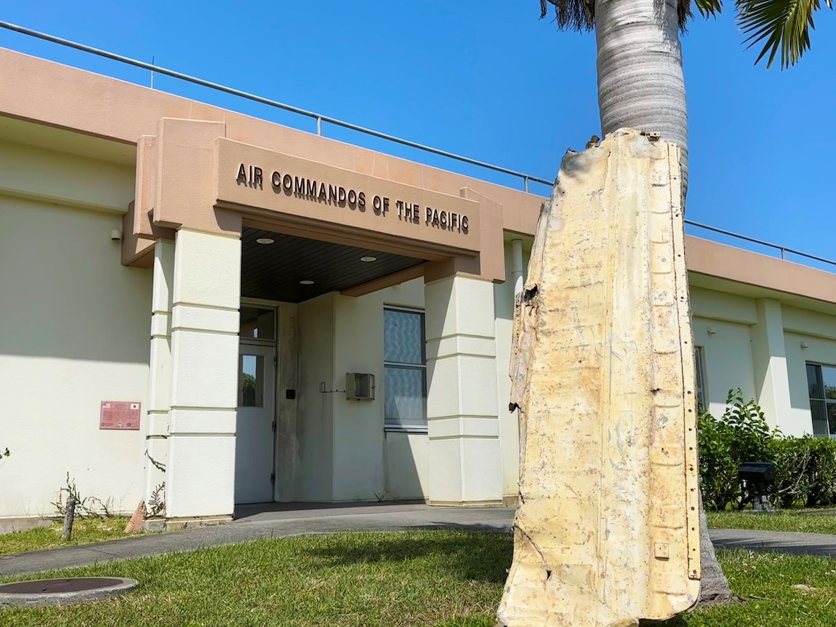 An old airplane wing rests against a tree in front of a building with the words "AIR COMMANDOS OF THE PACIFIC" above the door.