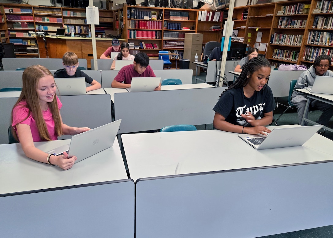 Students set a long tables in a classroom and work on laptops computers.