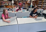Students set a long tables in a classroom and work on laptops computers.