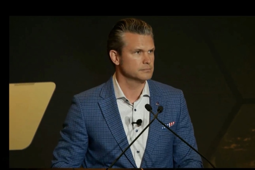 Defense Secretary Pete Hegseth stands in front of two microphones against a black background.